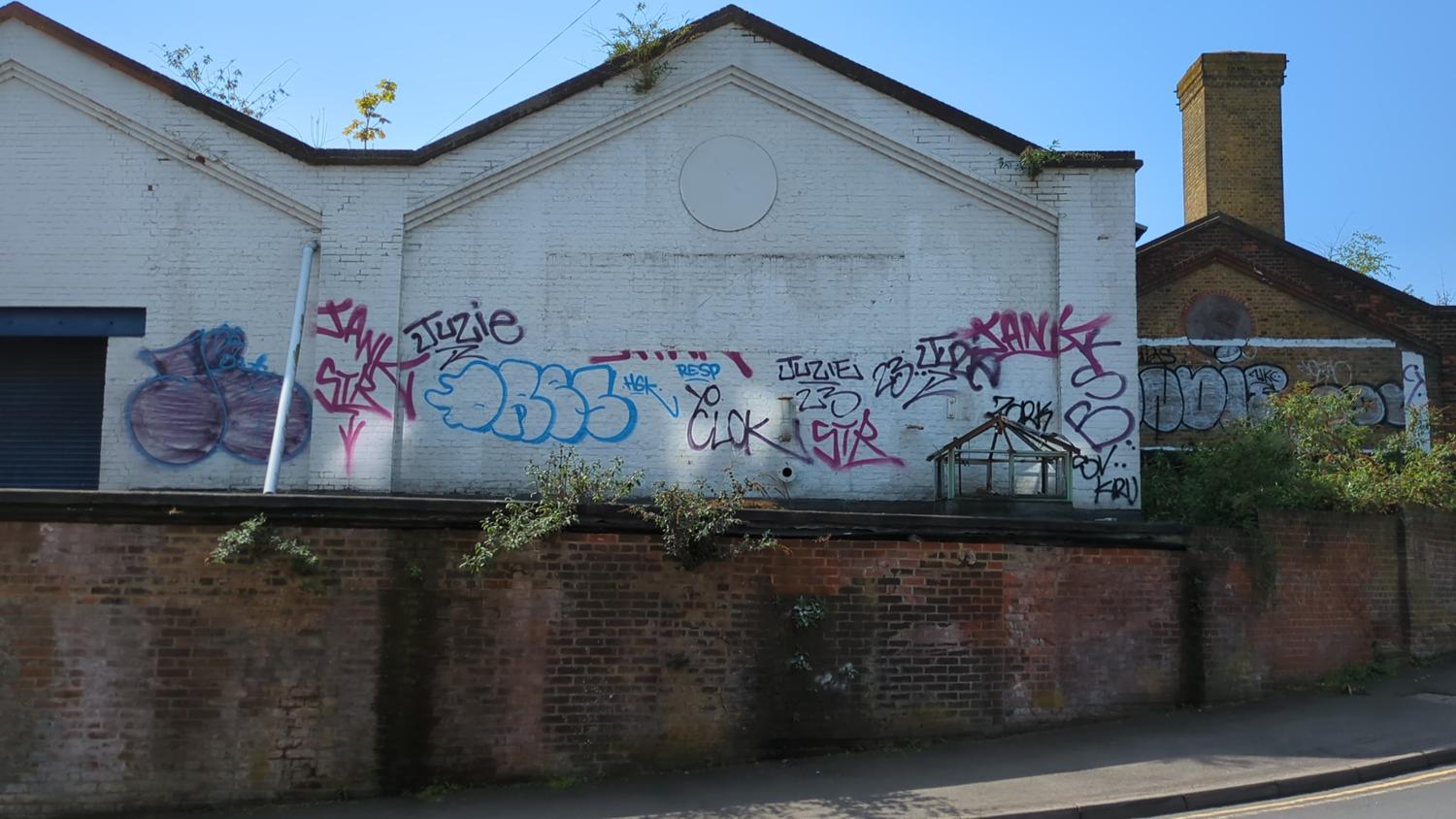 The image shows a section of an old brick and white brick building with graffiti on the walls. The graffiti is colorful and varied in style. Some plants are growing out of the brick wall and on the roof of the building. The building appears to be in a somewhat neglected state.