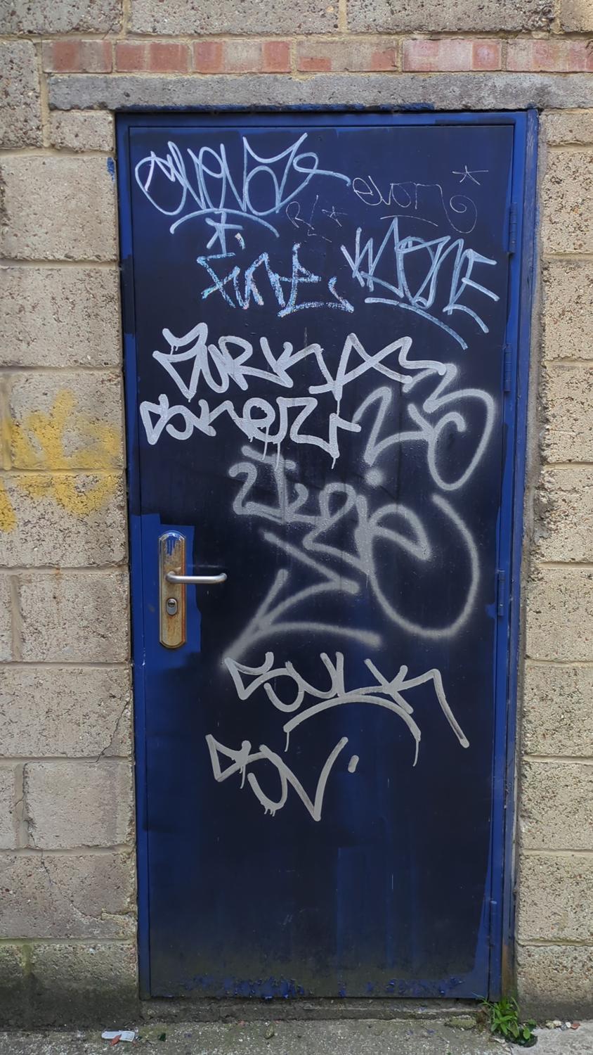 The image shows a blue metal door covered in white graffiti. The door is set into a brick wall. There is a simple metal handle on the door.