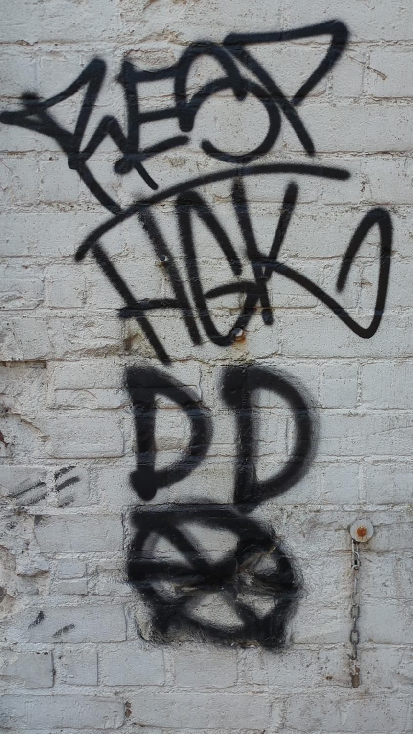 The image shows black spray paint graffiti on a white brick wall. The graffiti consists of abstract shapes and letter-like forms. A metal chain is attached to the wall near the graffiti.
