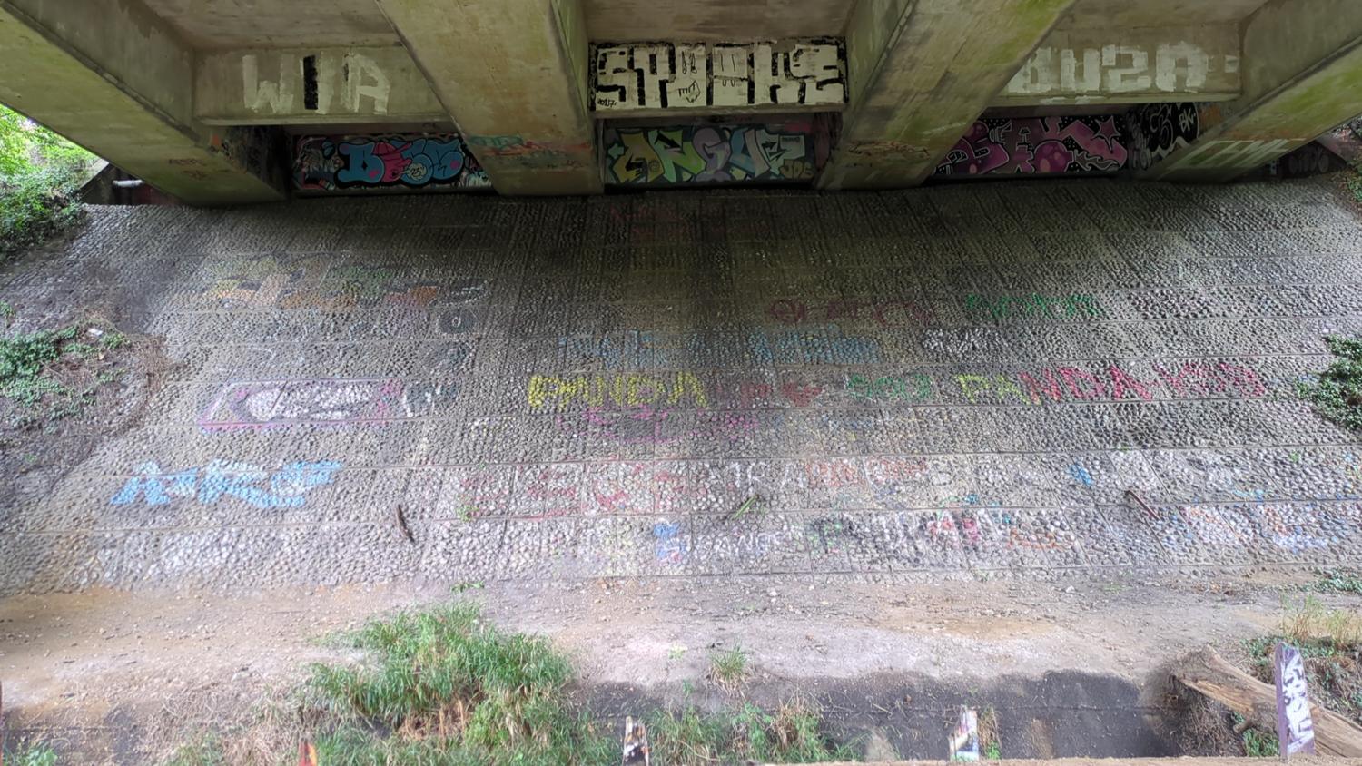 The image shows a concrete structure, possibly a bridge or overpass, with graffiti covering its surfaces. The structure is made of large, textured concrete blocks. Vegetation grows at the base of the structure. There is ground level at the base of the structure.