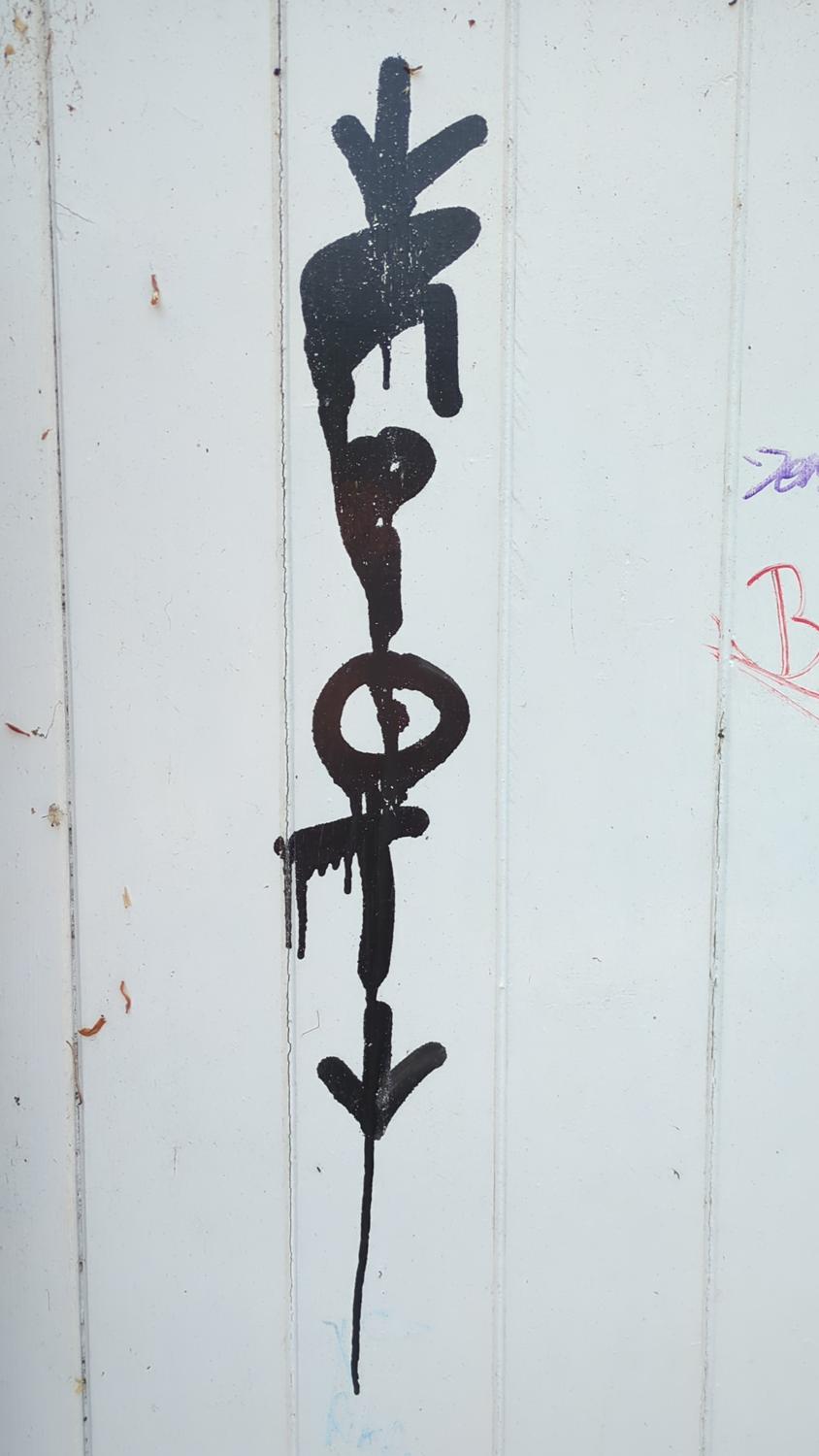 The image shows black graffiti on a white wooden surface. The graffiti appears to be an abstract design, with some resemblance to an arrow or stylized lettering.