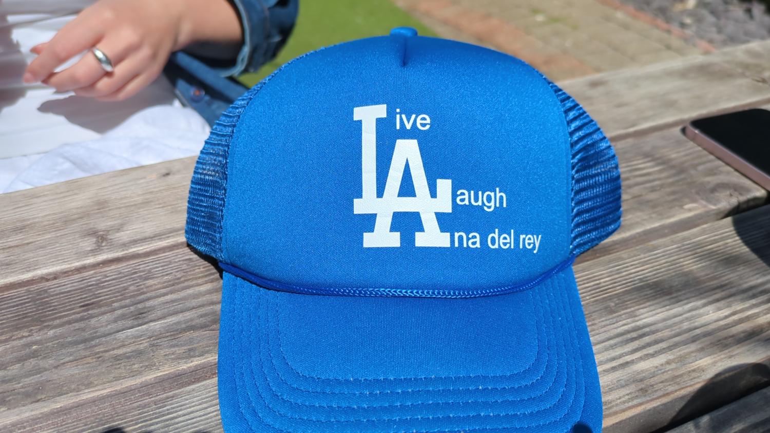 A bright blue baseball cap sits on a weathered wooden surface. The cap features a white design that incorporates the Los Angeles Dodgers 