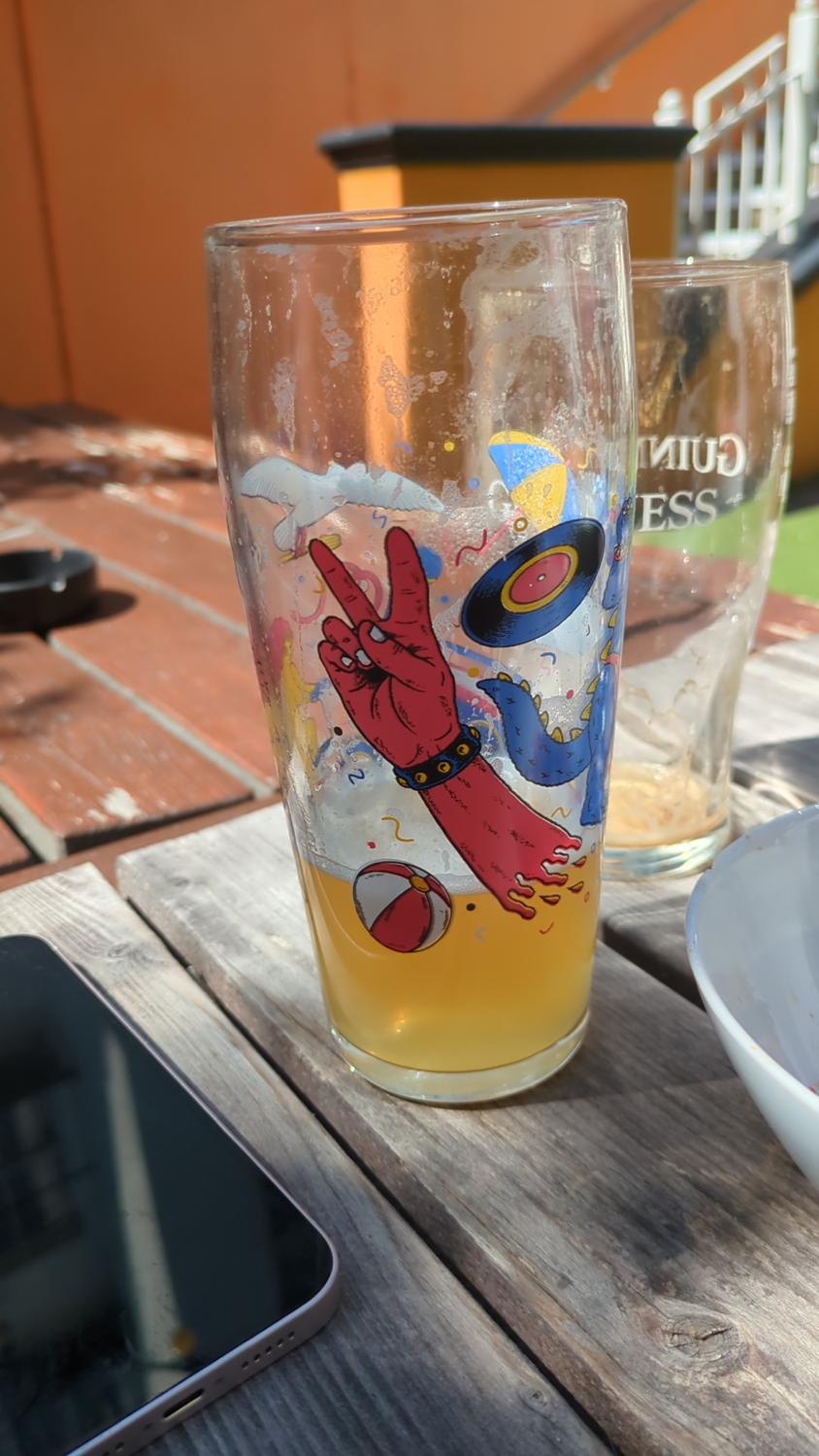 The image shows a partially full glass of beer with a colorful cartoon design on it, sitting on a wooden table. Another beer glass can be seen in the background. There is also a white bowl/container and a smartphone on the table.