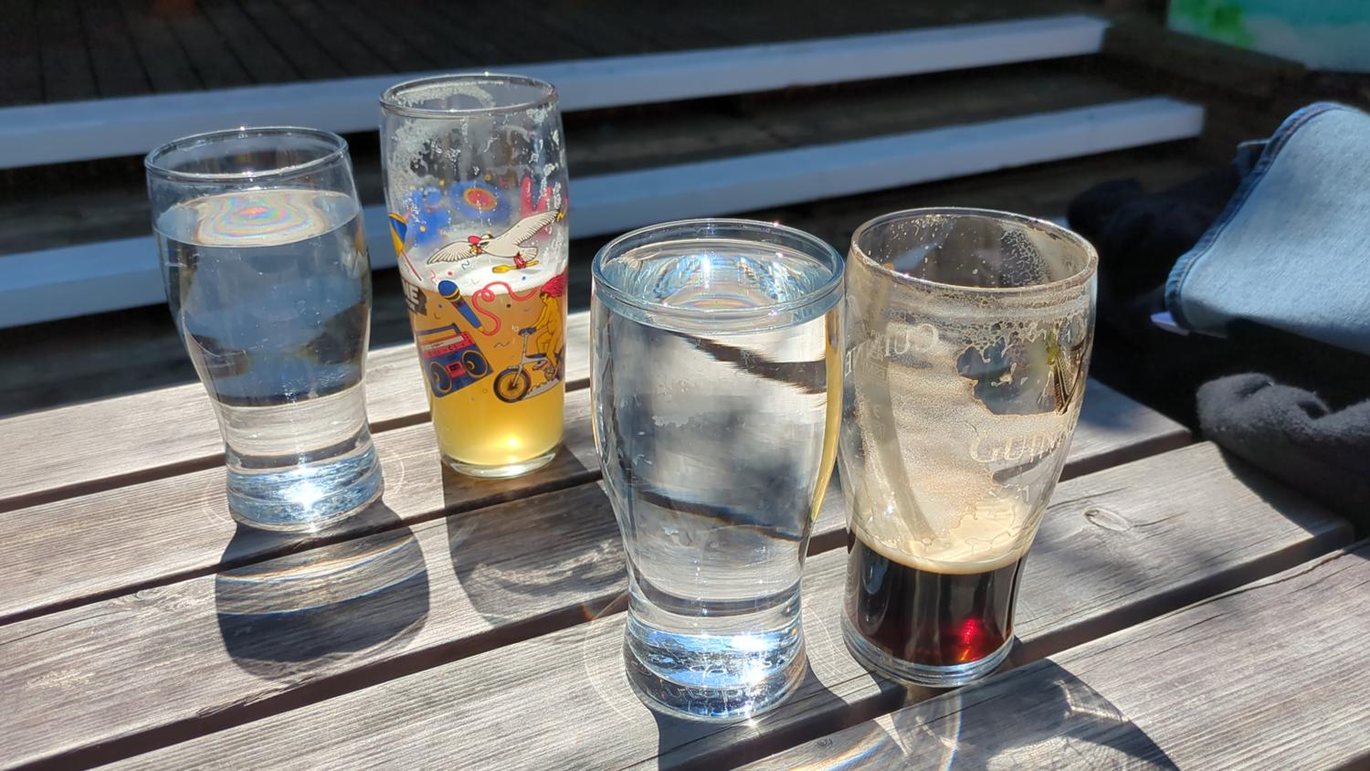The image shows four glasses on a wooden table. One glass is full of water, another contains a yellow liquid with a cartoon design, a third is filled with water, and the last appears to hold Guinness. Part of a leg wearing jeans and black clothing is also visible. The scene is outdoors, likely on a deck or patio.